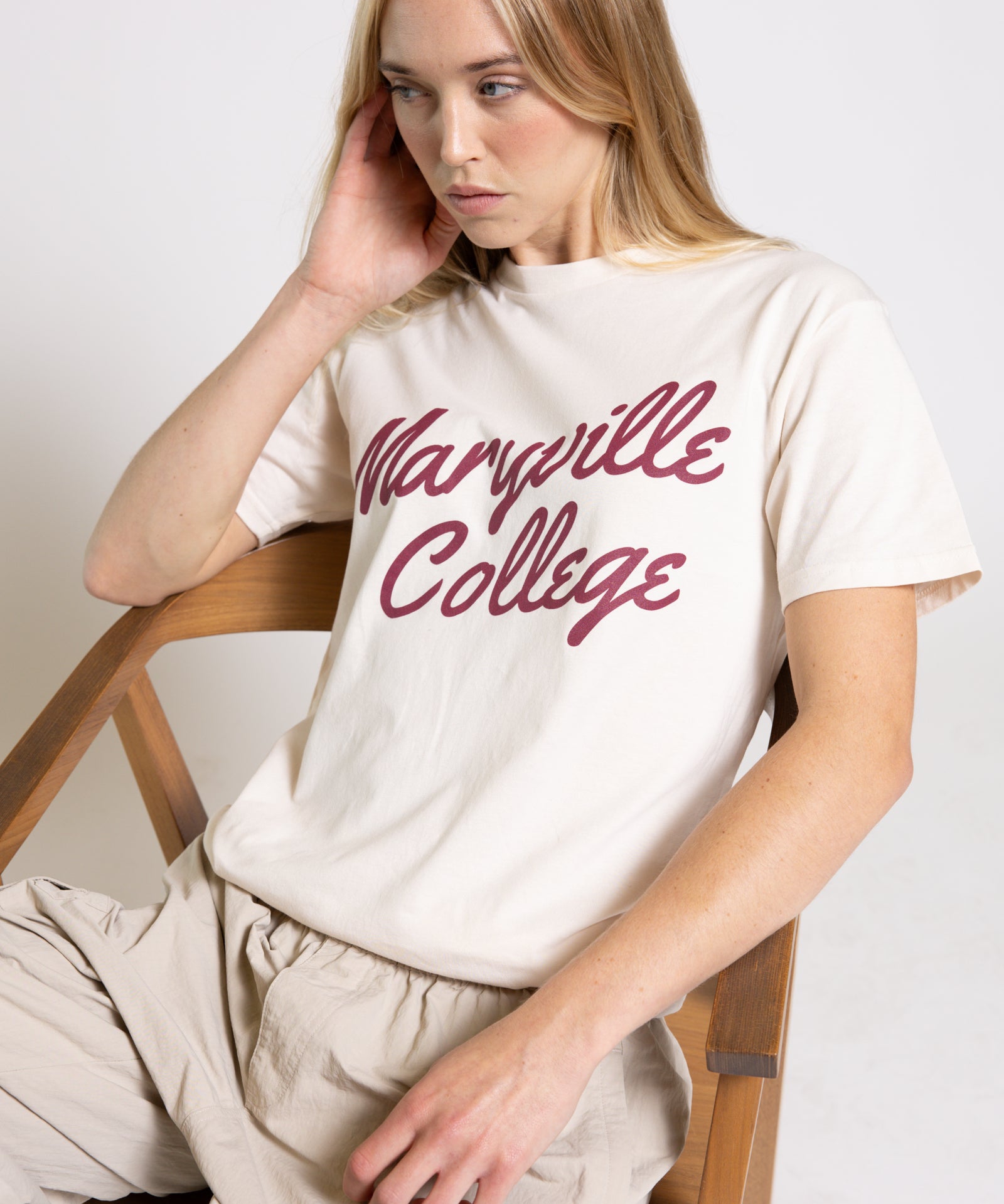 Woman wearing a cream t-shirt with 'Maryville College' script printed in red, sitting on a wooden chair.