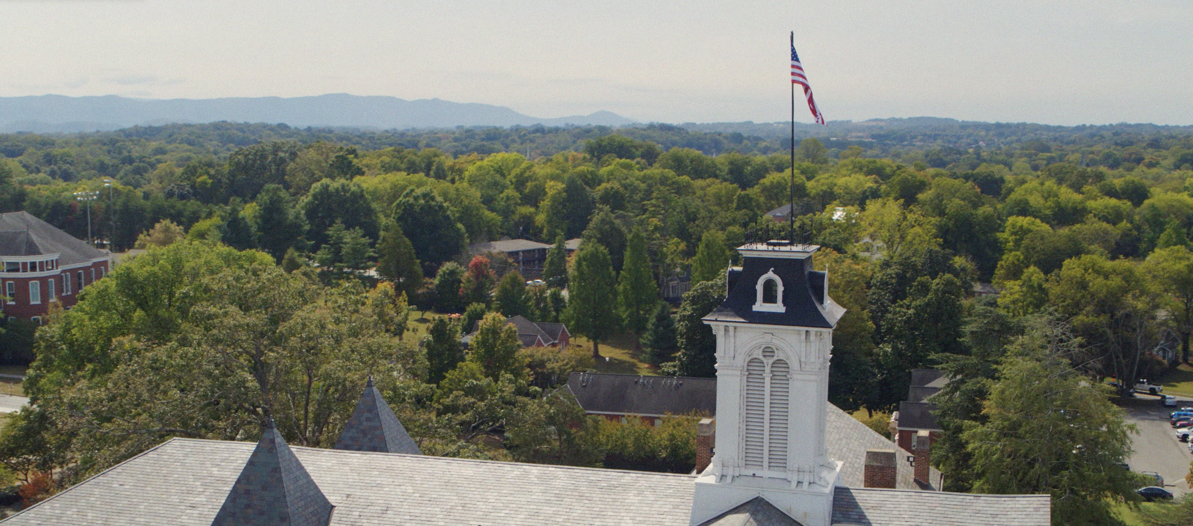 Aerial view of a Maryville College campus building with an American flag, surrounded by trees and rolling hills.